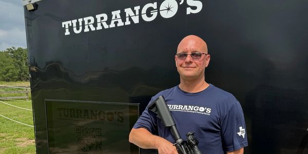 Picture of Founder Joe Turrigiano next to the Turrango's mobile trailer while holding a rifle