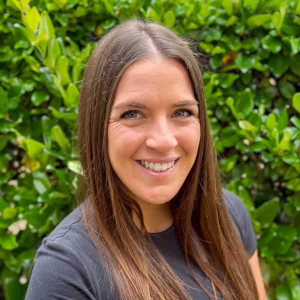 Smiling woman with long hair in front of green bushes.