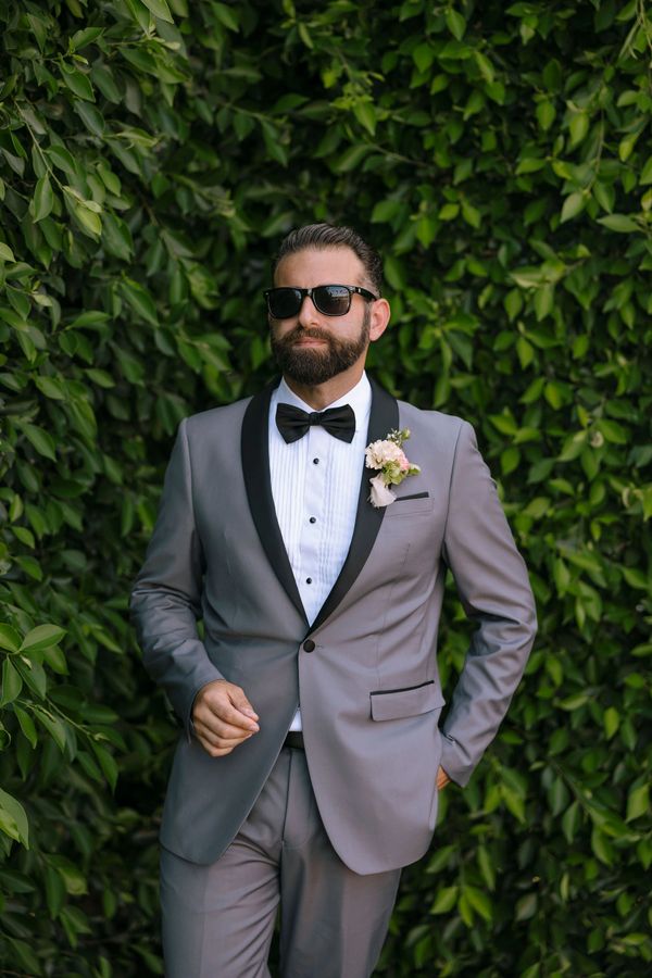 Man in a stylish grey tuxedo with a black bow tie and sunglasses, standing against a leafy green backdrop.