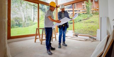 Two construction workers review blueprints inside a house with large windows overlooking greenery.