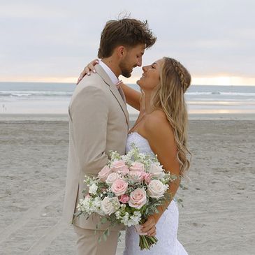 Newlyweds embrace on a beach at sunset, bride holding a bouquet of pink and white roses.