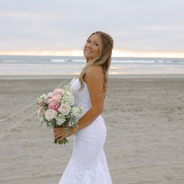 Bride in a white gown holding a bouquet on a beach at sunset.