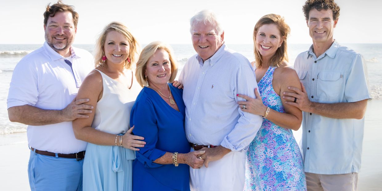 Six adults smiling at the beach in summer clothing.
