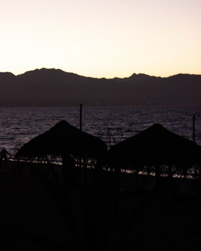 Silhouettes of beach umbrellas at dusk with mountains and calm sea.