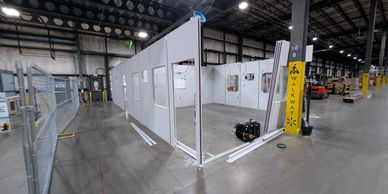 Modular office building under construction inside a large warehouse, with gray wall panels, door fra