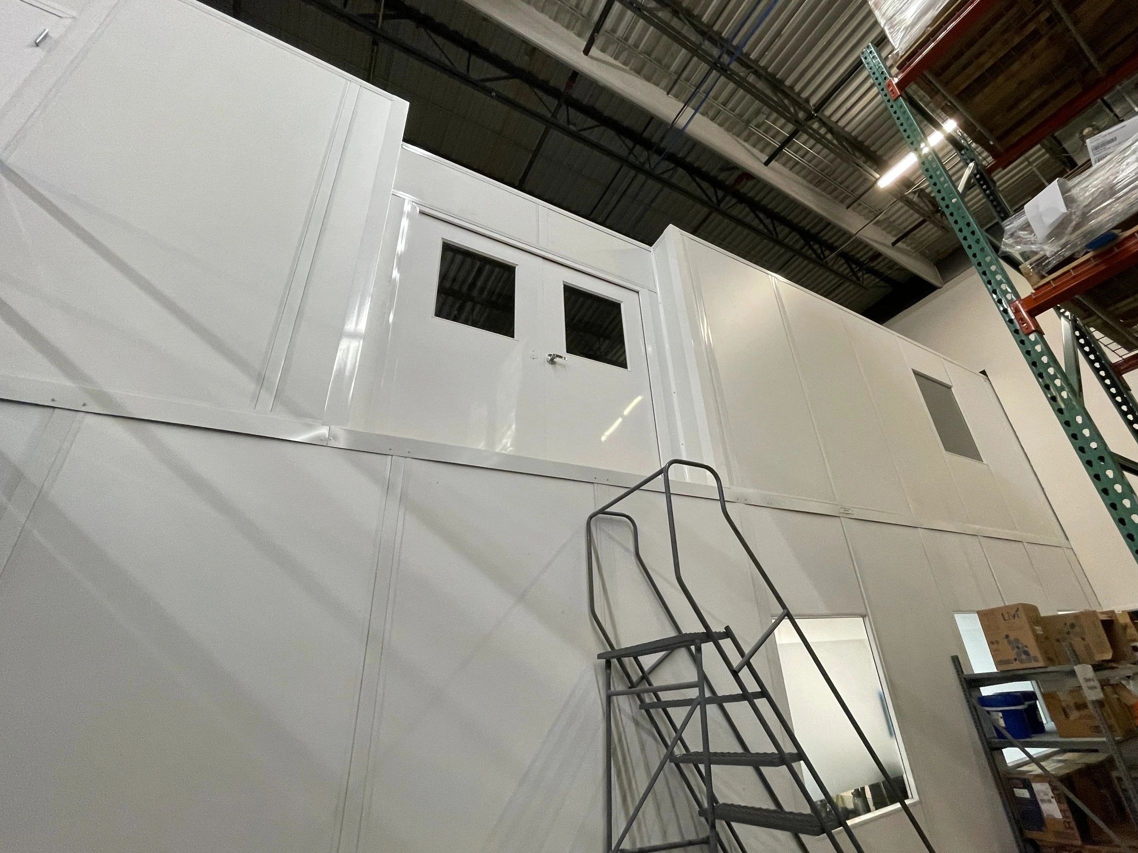 A two-story modular office building within a warehouse, featuring a double door and white walls.