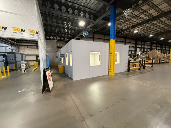A white modular office installation with multiple windows and a door, situated inside a warehouse
