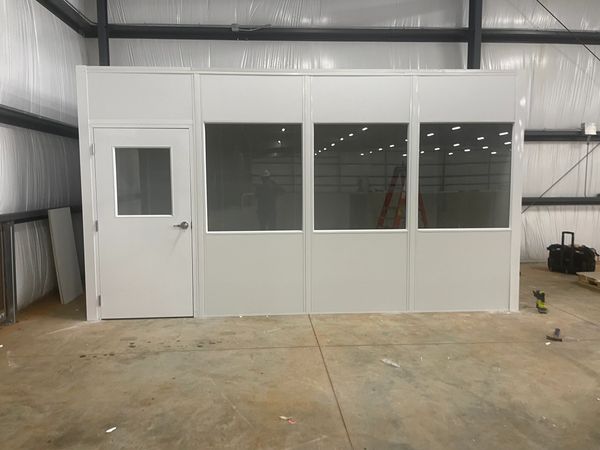 A prefabricated modular office with large windows and a solid door, installed inside a warehouse. 