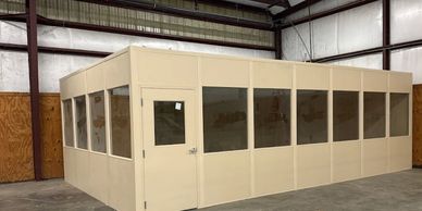A prefabricated modular in-plant office with beige walls and large windows, situated within a warehouse or industrial facility.
