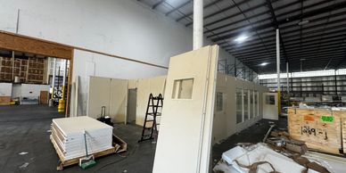A warehouse with modular office walls being constructed and building materials scattered on the floor.
