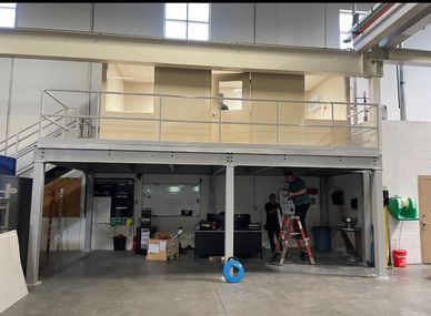 A beige modular office with several windows & a door sitting on top of a grey steel mezzanine & a se