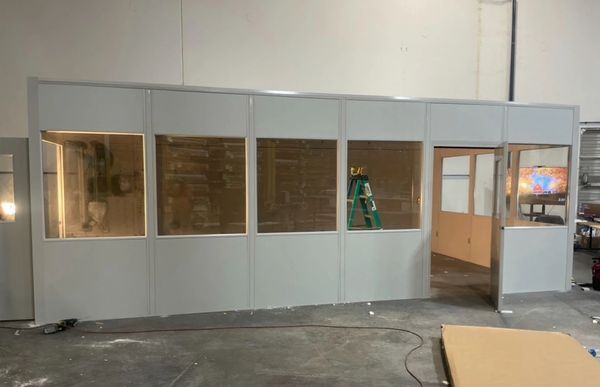 A modular clean-room with large windows and an open door, under construction in a warehouse.
