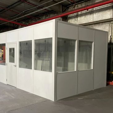 A prefabricated modular in-plant office with large windows and a white door, situated within a wareh