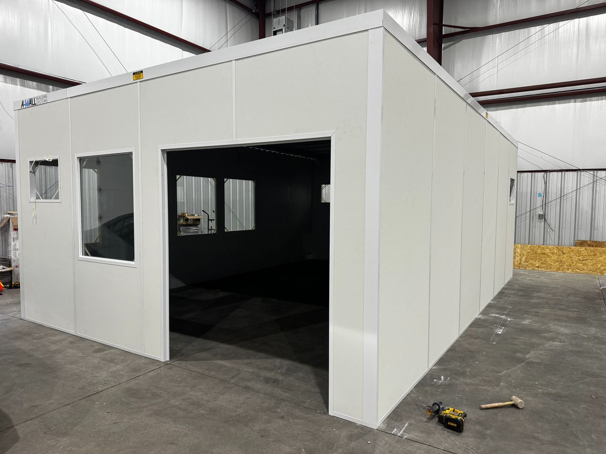 A prefabricated modular office with white walls & large windows, partially assembled in a warehouse.