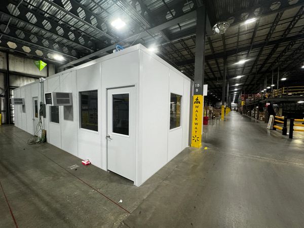 A white modular office installation with multiple windows and a door, situated inside a warehouse