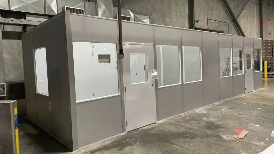 A modular office building with gray walls and multiple windows, situated in a warehouse setting."
