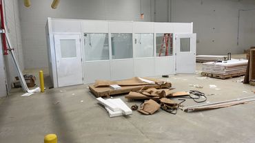 A modular office with white walls and large glass windows is being constructed inside a warehouse. 