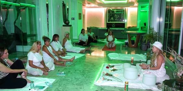 A group of women in white sitting on mats in a green-lit room, meditating or attending a spiritual session.