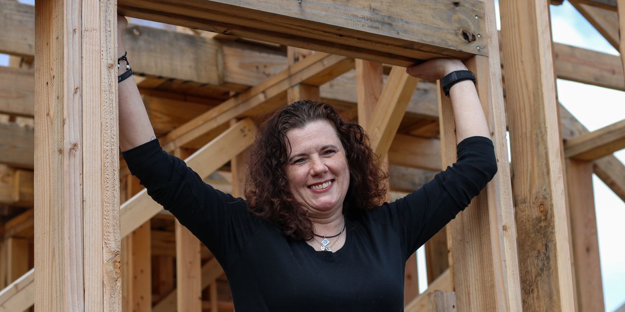Smiling woman holding onto wooden frame structure at construction site.