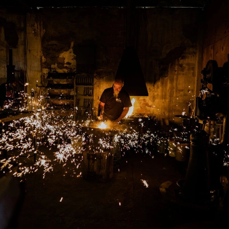 A blacksmith works on metal, sparks flying in a dimly lit workshop.