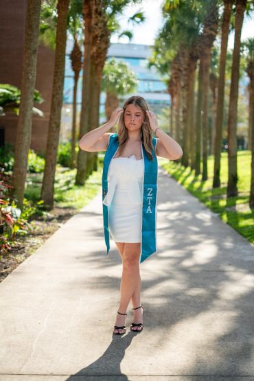 Young woman in white dress and ZTA stole walks on sunny pathway lined with palm trees.