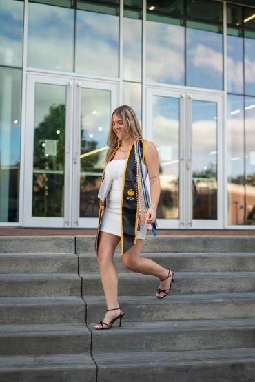 Young woman in graduation attire walking down steps outside a modern building.