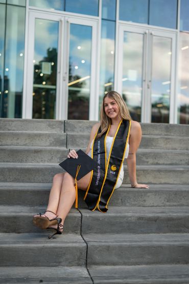 Young woman in graduation attire sitting on steps outside a building.