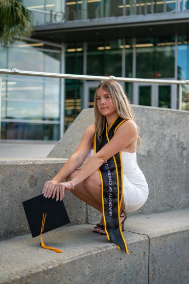 A young woman in a white dress with a graduation stole, holding a cap.