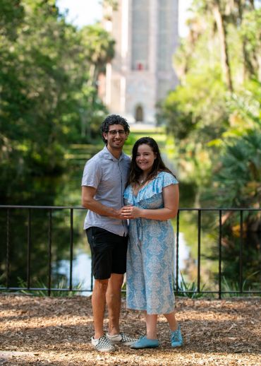 Couple standing by a railing near a calm water body surrounded by greenery.