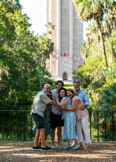 A happy family group hugging outdoors near a tall tower surrounded by greenery.
