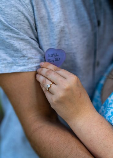 A hand with an engagement ring holds a heart-shaped note saying 'Will You Marry Me?'