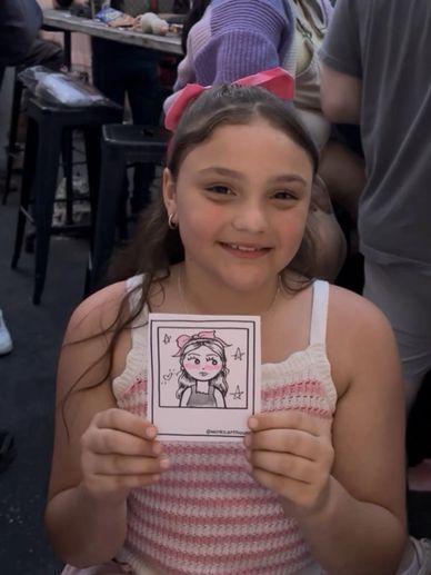 Smiling girl holding a cartoon portrait of herself at an outdoor event.