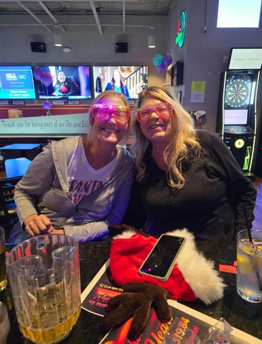 Two women wearing festive light-up glasses smiling in a bar.