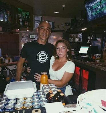 Two people smiling behind a bar counter with drinks and food.