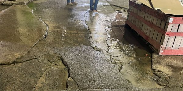 Two men stand on a cracked, wet concrete floor near a large wooden crate indoors.
