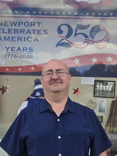 Man in blue shirt stands in front of a patriotic Newport 250th anniversary banner.