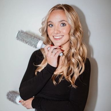 Smiling woman with long blonde hair holding round hairbrushes.