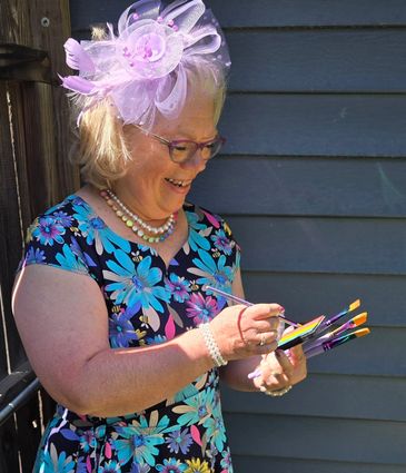 Smiling woman painting outdoors in a floral dress and pink fascinator.