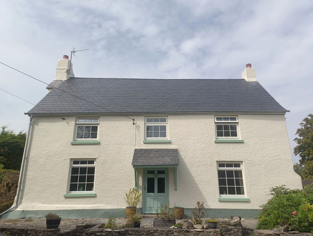 White cottage with green trims and slate roof under a cloudy sky.