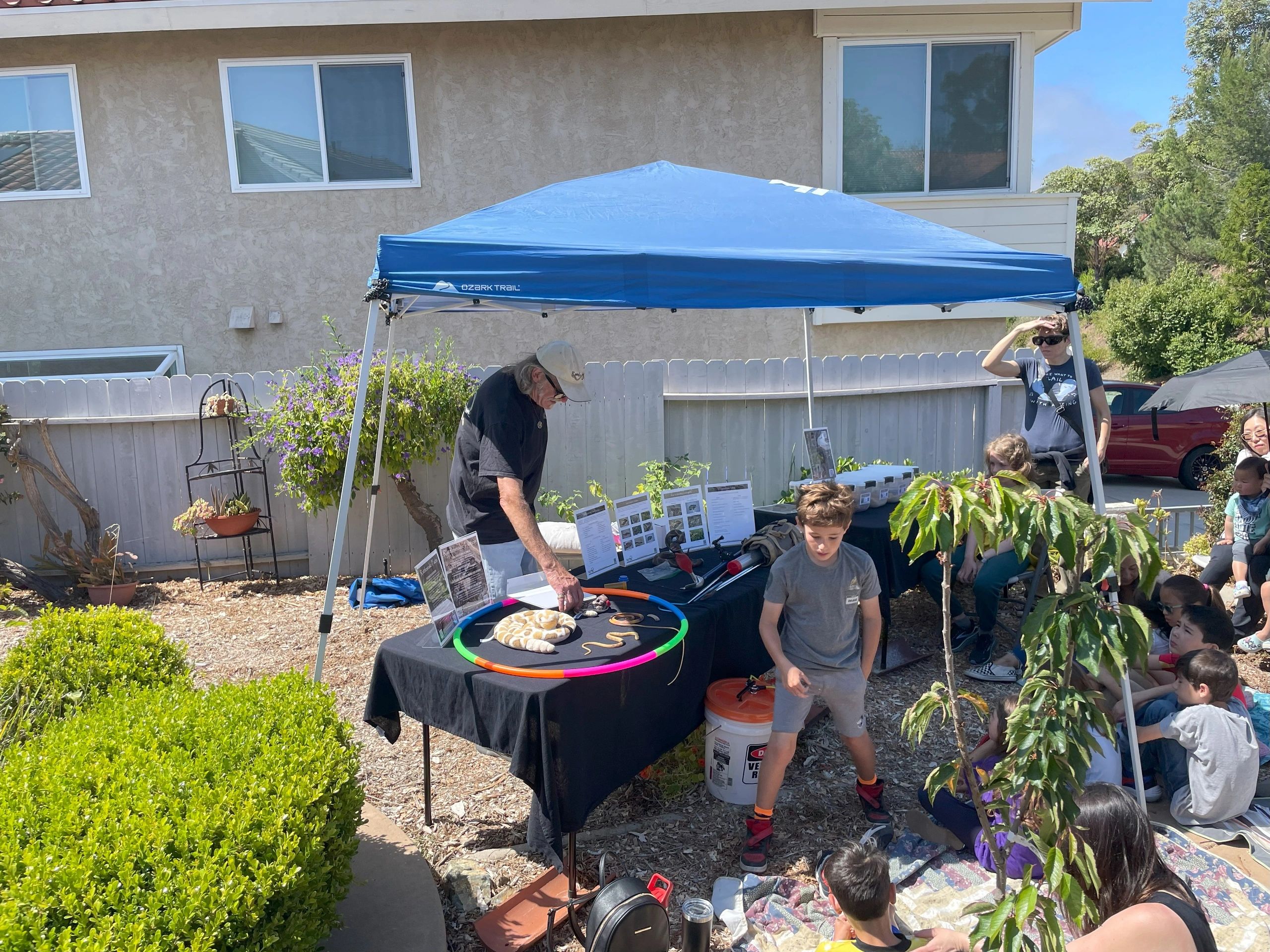Outdoor educational setup with people, a blue canopy, and a table displaying a snake.
