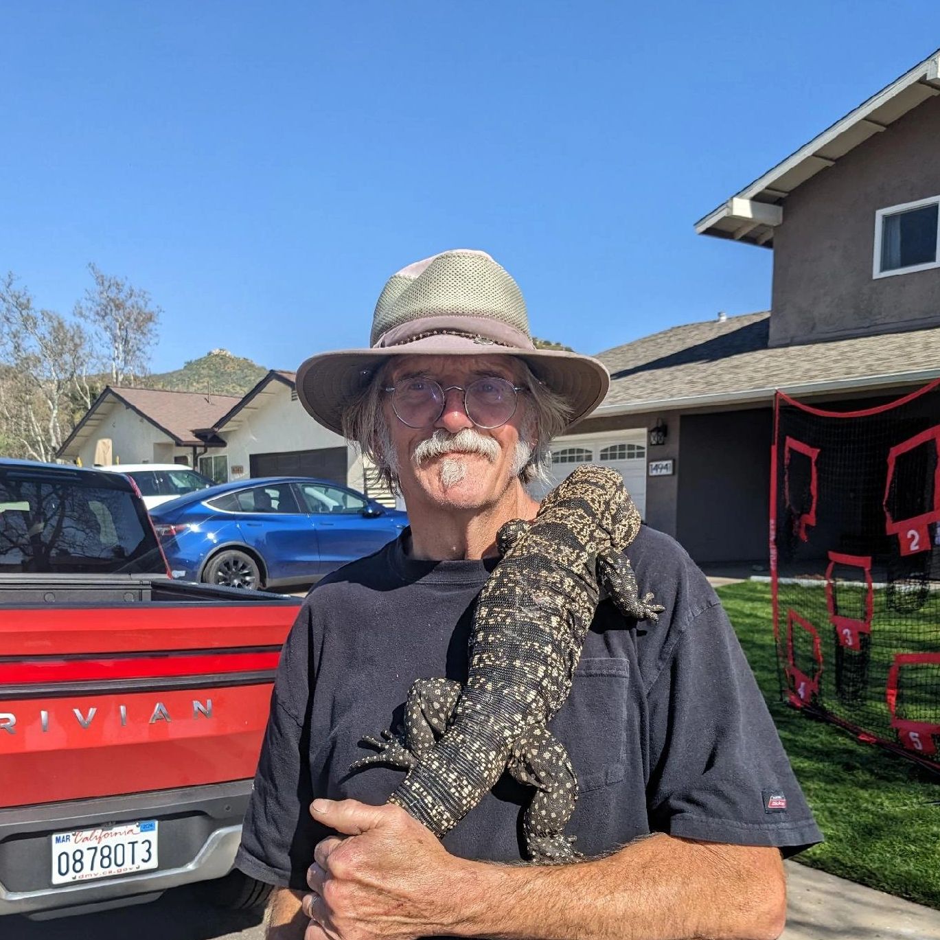Man with glasses and hat holding a large lizard outdoors.