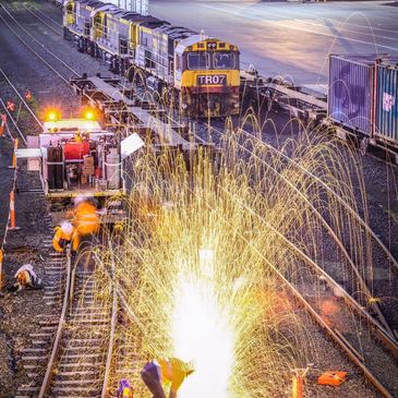 Welding sparks illuminate workers on train tracks at dusk, with a train and shipping containers 