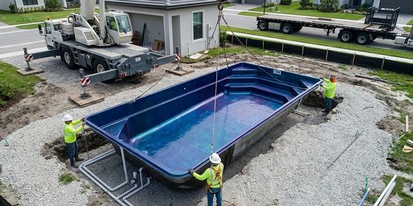 Workers install a large blue fiberglass pool in a backyard under construction.
