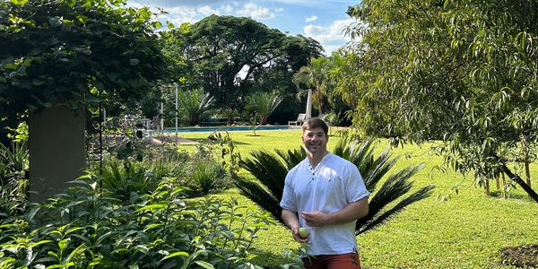 Man smiling in a lush garden holding a tennis ball under bright sunlight.