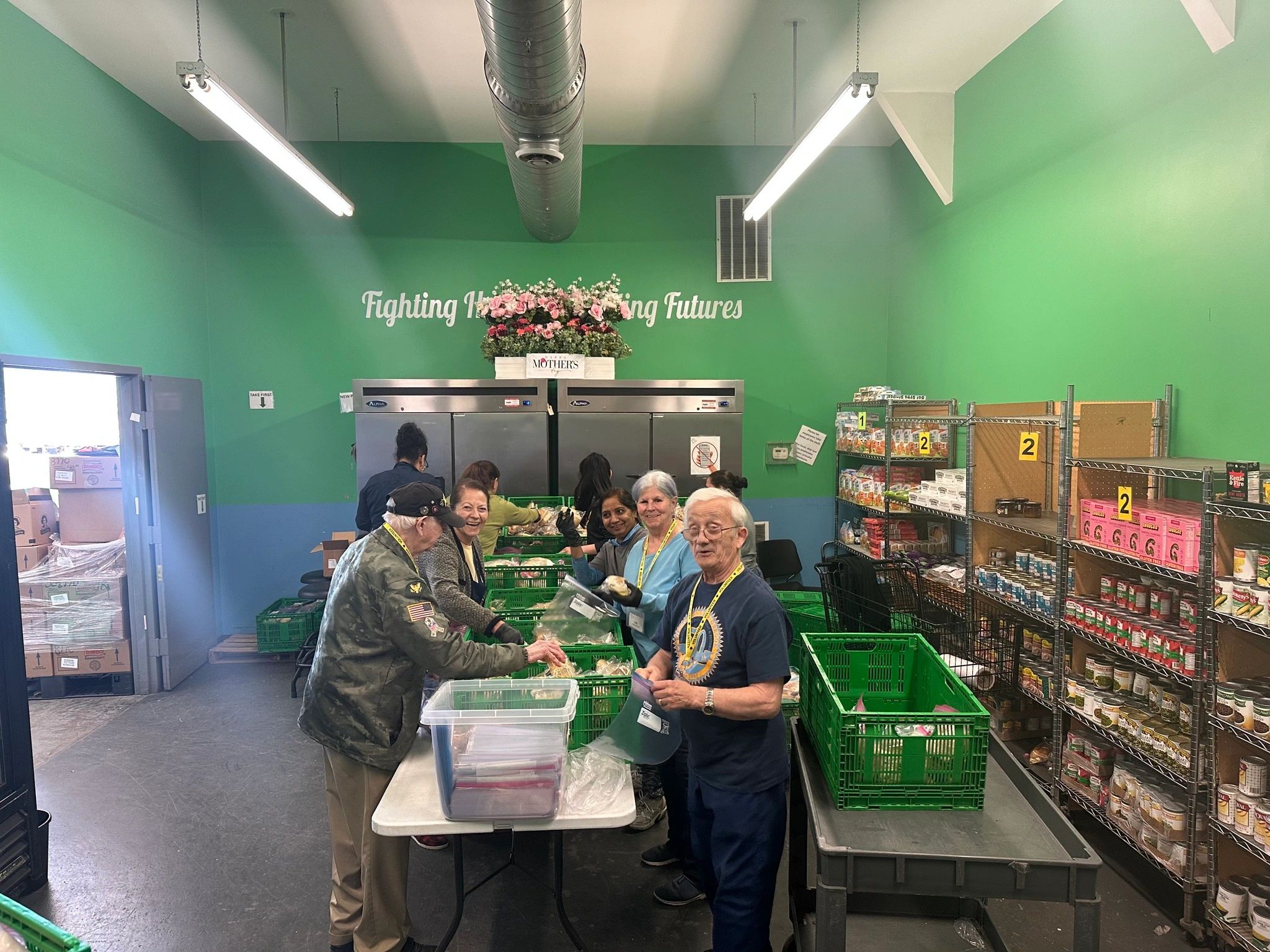 Volunteers packing food at a community center with green walls and shelves of supplies.