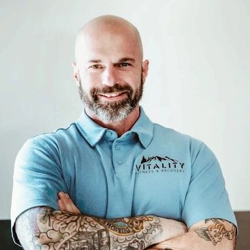 Smiling bald man with beard and tattoos in a blue Vitality polo shirt.