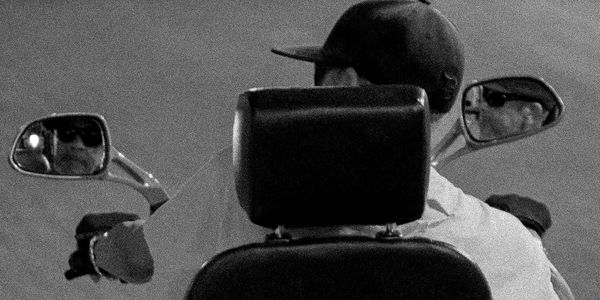 A black and white photo of a man on a mobility scooter from behind looking in his rear-view mirror