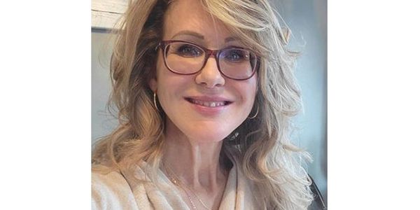 Smiling woman with blonde hair and glasses in a cozy setting.