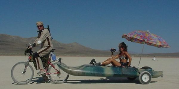 Person on bike towing a canoe with a woman and umbrella in a desert.