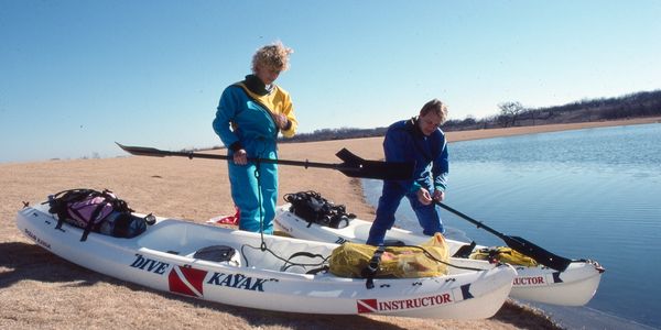Two people preparing kayaks labeled 'Instructor' by a calm lake shore.